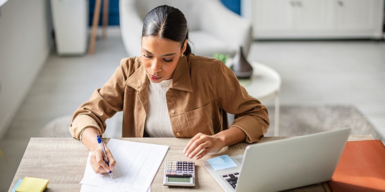 A young hispanic woman is focused on preparing her taxes at a home office. She writes notes, uses a calculator, and organizes her financial documents on the desk.