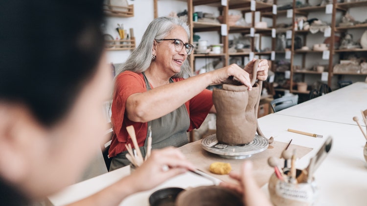 A woman with gray hair shapes a piece of clay on a pottery wheel