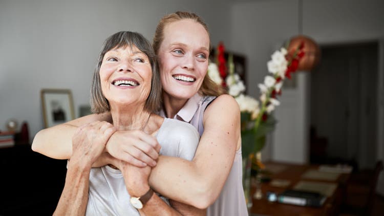 A young woman hugs an older woman from behind, both smiling