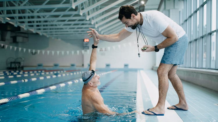 A swimming coach high-fiving an elderly male swimmer over the edge of an indoor pool