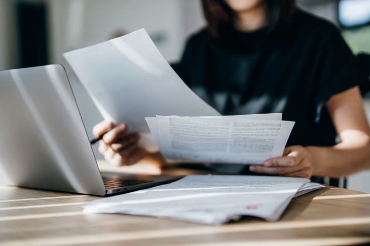 A person sits at a desk holding papers and working on a laptop